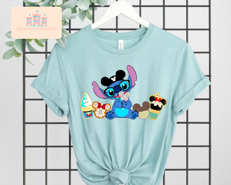 Puede incluir: Una camiseta azul claro con un personaje Stitch azul que lleva orejas y gafas de Mickey Mouse, sosteniendo una piruleta. Stitch est&aacute; rodeado de varias golosinas con tem&aacute;tica de Disney, incluyendo un cono de helado de Mickey Mouse, una dona de Minnie Mouse y un pretzel de Mickey Mouse.