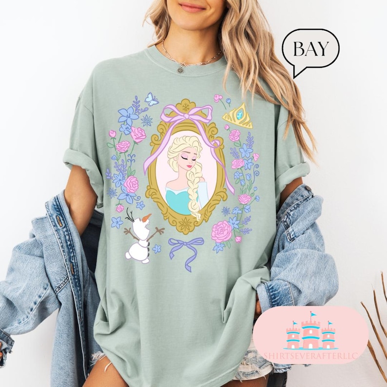 K&ouml;nnte beinhalten: Ein hellgr&uuml;nes T-Shirt mit einem floralen Design, das ein gerahmtes Bild von Elsa aus Frozen zeigt. Der Rahmen ist golden mit einem blauen und wei&szlig;en Band. Ein kleiner Schneemann befindet sich in der linken unteren Ecke des Designs.
