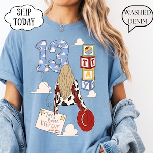 May include: A light blue t-shirt featuring a graphic of a person with long blonde hair, a cow-print jacket with red fringe, and a red hat. The design includes a number 13 balloon, alphabet blocks spelling TAY, and a sign reading TAY'S ROOM KEEP OUT.