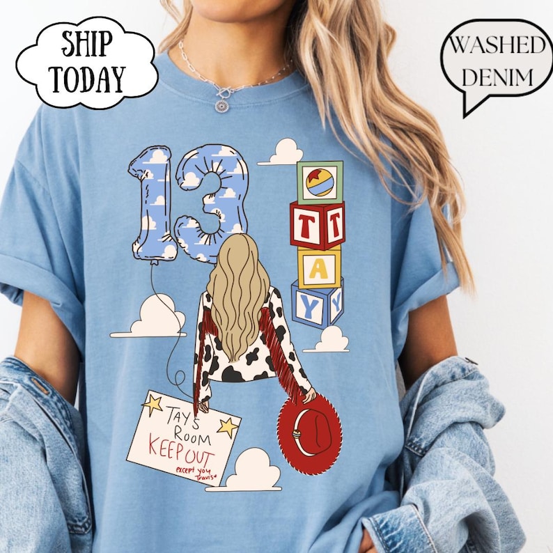 May include: A light blue t-shirt featuring a graphic of a person with long blonde hair, a cow-print jacket with red fringe, and a red hat. The design includes a number 13 balloon, alphabet blocks spelling TAY, and a sign reading TAY'S ROOM KEEP OUT.