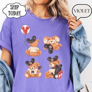 May include: A violet t-shirt featuring a graphic print of four brown teddy bears wearing black mouse ear hats. The bears are depicted in various poses, including one holding a red balloon, one upside down, one with a letter M shirt, and one with a backpack.
