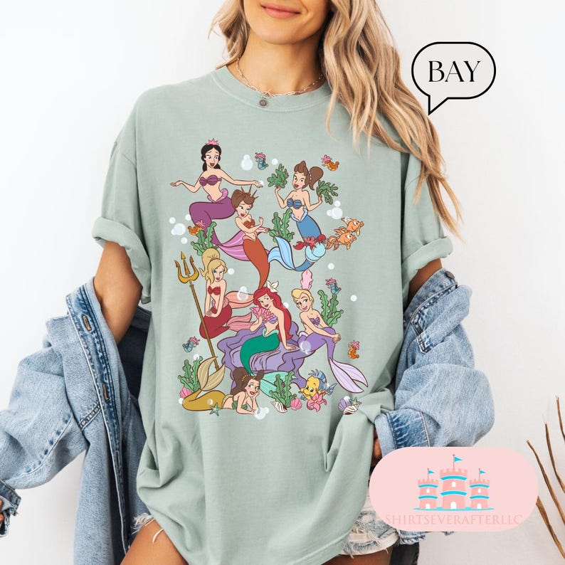 May include: A light green t-shirt featuring a colorful illustration of mermaids in a sea scene. The graphic includes various mermaids with different hair colors and tail designs, surrounded by aquatic plants and sea creatures. The word "BAY" is in a speech bubble.