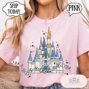 May include: Pink t-shirt featuring a whimsical castle design with blue turrets, gold accents, and the words "Disney World." The shirt includes the text "Ship Today" and "Pink."