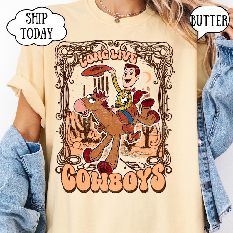 May include: A light yellow t-shirt featuring a cartoon illustration of Woody from Toy Story riding a horse. The graphic includes the words "Long Live Cowboys" in a vintage style. The shirt has a crew neck and short sleeves.