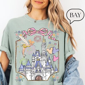 Puede incluir: Camiseta verde claro con una ilustraci&oacute;n caprichosa de un hada, un castillo, fuegos artificiales y globos de Mickey Mouse. El dise&ntilde;o est&aacute; enmarcado por un lazo rosa y detalles florales. La palabra "BAY" est&aacute; en una burbuja.