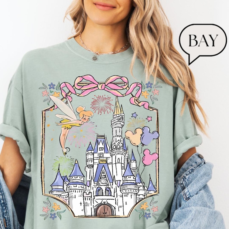 Puede incluir: Camiseta verde claro con una ilustraci&oacute;n caprichosa de un hada, un castillo, fuegos artificiales y globos de Mickey Mouse. El dise&ntilde;o est&aacute; enmarcado por un lazo rosa y detalles florales. La palabra "BAY" est&aacute; en una burbuja.