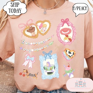 Op de afbeelding: Een perzikkleurig T-shirt met illustraties van stripfiguren in kaders en een banner met de tekst "TO INFINITY AND BEYOND". Het ontwerp bevat een hartvormig kader met een cowboy en een prinses, en een Buzz Lightyear-afbeelding.