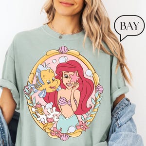 May include: A light green t-shirt featuring a cartoon illustration of Ariel from The Little Mermaid. Ariel has red hair and is holding a pink starfish. Flounder is next to her. The word "BAY" is in a speech bubble.