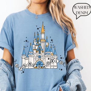 May include: Light blue t-shirt featuring a detailed illustration of a fairytale castle with blue turrets, gold accents, and the text "Disney World" in a circular design. The shirt has a relaxed fit and the words "Washed Denim" in a speech bubble.