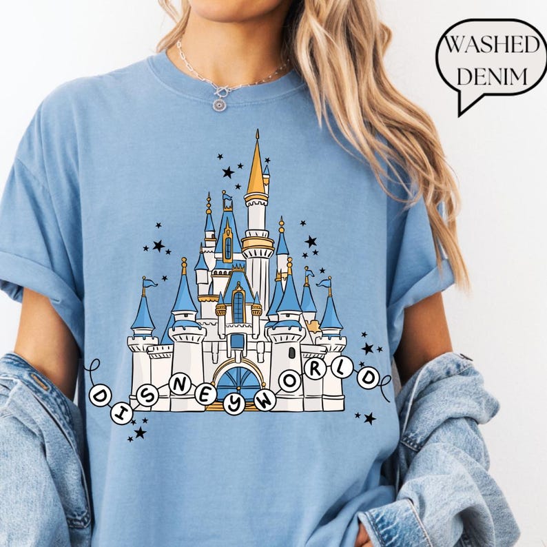 May include: Light blue t-shirt featuring a detailed illustration of a fairytale castle with blue turrets, gold accents, and the text "Disney World" in a circular design. The shirt has a relaxed fit and the words "Washed Denim" in a speech bubble.