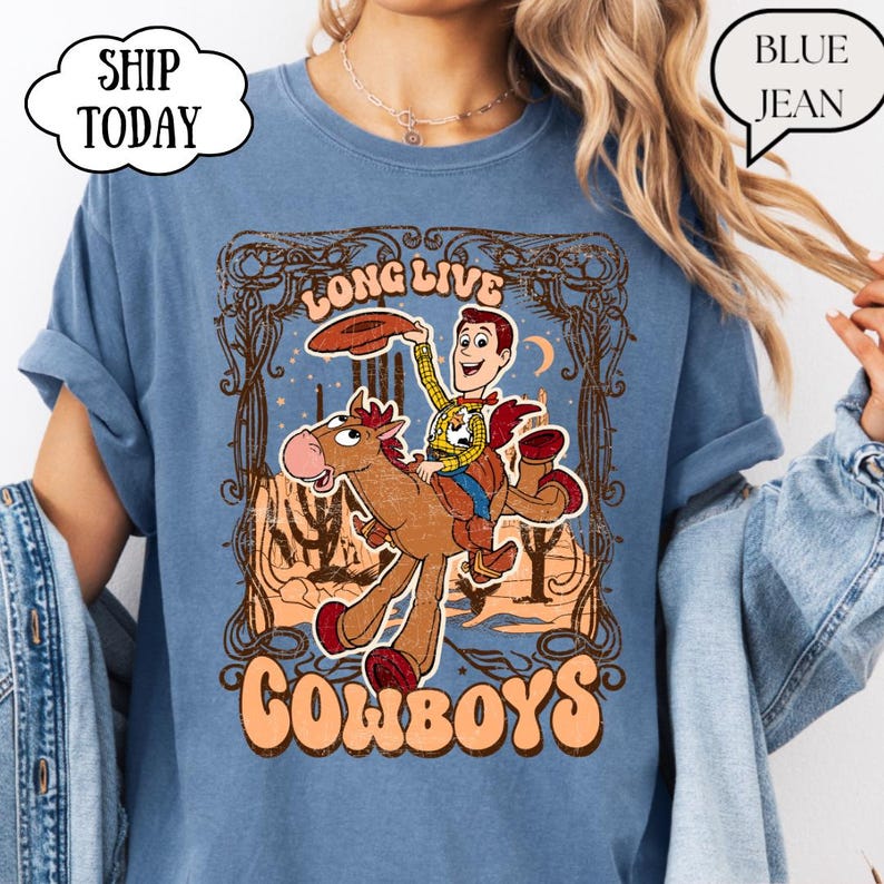 May include: A blue t-shirt featuring a cartoon cowboy riding a horse, with the words "LONG LIVE COWBOYS" in an orange font. The design includes a cowboy hat and a desert landscape. The shirt has rolled-up sleeves.