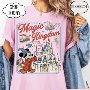 May include: Pink t-shirt with a graphic featuring Mickey Mouse as a wizard, a castle, and the words "Magic Kingdom." The design includes images of Dumbo, a trolley car, and Tinkerbell. The shirt has a blue border and star accents.