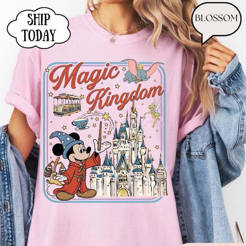 May include: Pink t-shirt with a graphic featuring Mickey Mouse as a wizard, a castle, and the words "Magic Kingdom." The design includes images of Dumbo, a trolley car, and Tinkerbell. The shirt has a blue border and star accents.