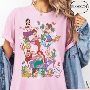 Op de afbeelding: Lichtroze T-shirt met een kleurrijke illustratie van verschillende zeemeerminnen met verschillende haarkleuren en zeemeerminstaarten. Het ontwerp bevat zeedieren en zeewier. Het woord "BLOSSOM" staat in een tekstballon.