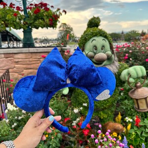 May include: Blue velvet Minnie Mouse ears with a sparkly heart charm. The ears are held in front of a topiary of a Disney character.