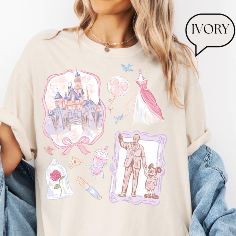 Op de afbeelding: Ivoor kleurig t-shirt met diverse roze en blauwe illustraties, waaronder een kasteel, een jurk en een ingelijste afbeelding van een man en een cartoonmuis. Het woord "IVORY" staat in een tekstballon.