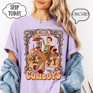 May include: A light purple t-shirt featuring a cartoon cowboy riding a horse, with the words "LONG LIVE COWBOYS" in a decorative frame. The cowboy is holding a hat, and the design has a vintage, western theme.