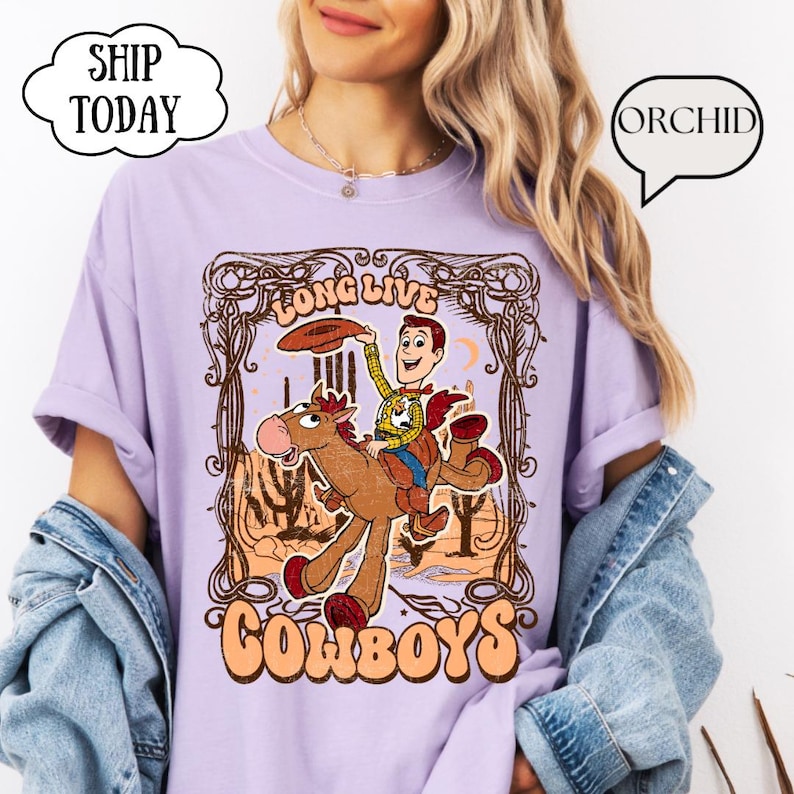 May include: A light purple t-shirt featuring a cartoon cowboy riding a horse, with the words "LONG LIVE COWBOYS" in a decorative frame. The cowboy is holding a hat, and the design has a vintage, western theme.