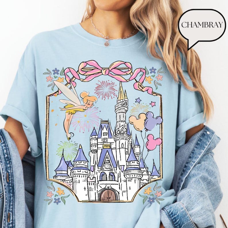 Puede incluir: Camiseta azul claro con un gr&aacute;fico de un castillo de cuento de hadas, Campanilla, fuegos artificiales y globos de Mickey Mouse. El dise&ntilde;o est&aacute; enmarcado con un lazo rosa y detalles florales. La palabra "CHAMBRAY" est&aacute; en una burbuja.