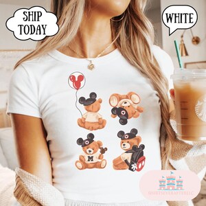 May include: A white t-shirt featuring a graphic print of four brown teddy bears wearing black mouse ear hats. One bear holds a red balloon, another is upside down, one holds a small treat, and the fourth wears a backpack with red accents.