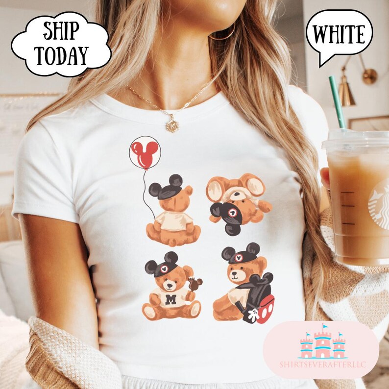 May include: A white t-shirt featuring a graphic print of four brown teddy bears wearing black mouse ear hats. One bear holds a red balloon, another is upside down, one holds a small treat, and the fourth wears a backpack with red accents.