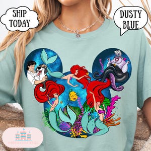 Puede incluir: Camiseta azul polvoriento con un dise&ntilde;o inspirado en Disney con Ariel y otros personajes de La Sirenita. El dise&ntilde;o incluye el texto "Dusty Blue" en una burbuja de di&aacute;logo. La camiseta tambi&eacute;n tiene el texto "Ship Today" en una burbuja de di&aacute;logo.