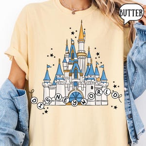 May include: A light yellow t-shirt featuring a detailed illustration of a castle with blue turrets and gold accents. The text "DISNEY WORLD" is arched below the castle, surrounded by stars.