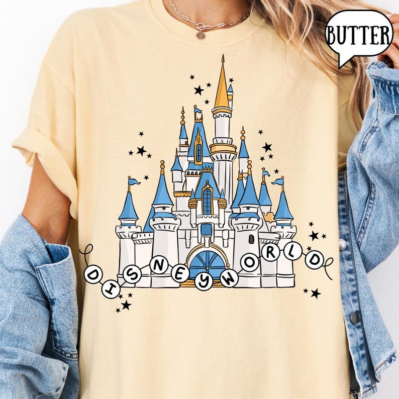 May include: A light yellow t-shirt featuring a detailed illustration of a castle with blue turrets and gold accents. The text "DISNEY WORLD" is arched below the castle, surrounded by stars.