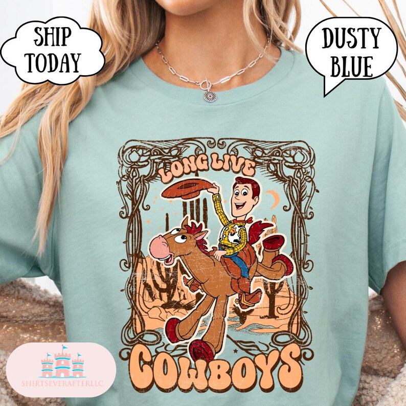 May include: Dusty blue t-shirt featuring a cartoon cowboy riding a horse, with the words "Long Live Cowboys" in a vintage style. The shirt has a distressed graphic design and the text "Dusty Blue" in a speech bubble.