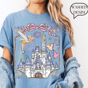 Puede incluir: Camiseta azul claro con un gr&aacute;fico de un hada, un castillo, fuegos artificiales y globos de Mickey Mouse. El dise&ntilde;o est&aacute; enmarcado por un lazo rosa y detalles florales. El texto "Washed Denim" est&aacute; en una burbuja.