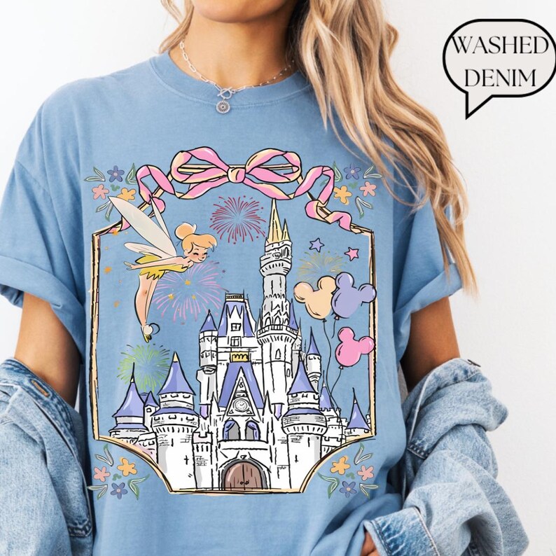 Puede incluir: Camiseta azul claro con un gr&aacute;fico de un hada, un castillo, fuegos artificiales y globos de Mickey Mouse. El dise&ntilde;o est&aacute; enmarcado por un lazo rosa y detalles florales. El texto "Washed Denim" est&aacute; en una burbuja.