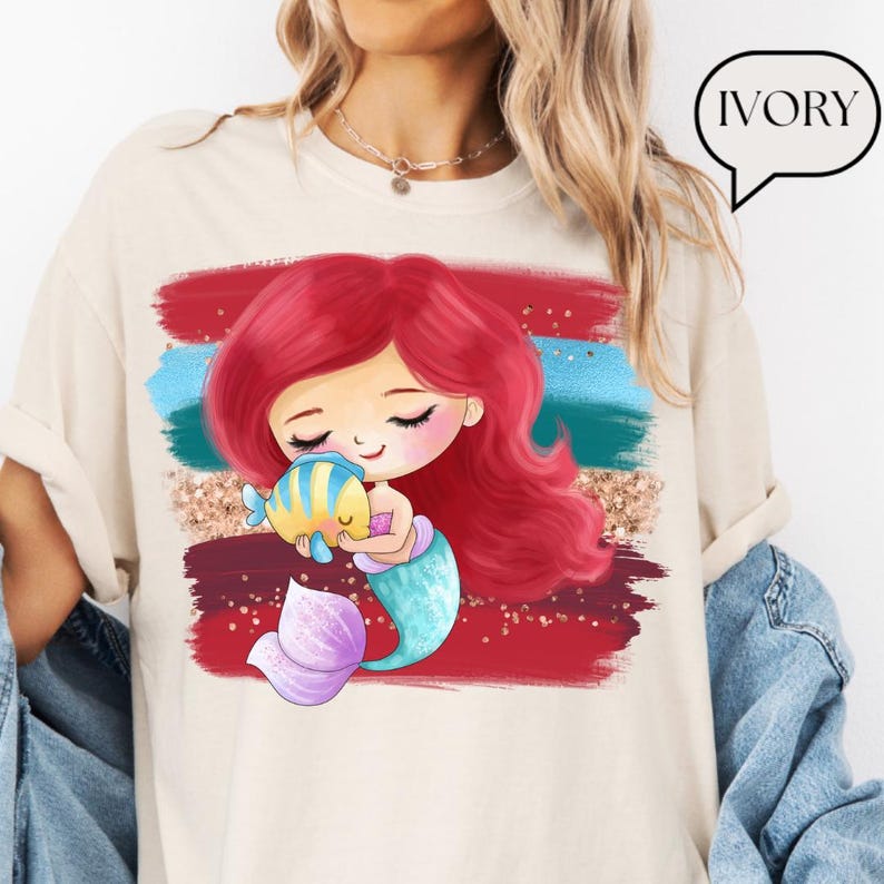 Puede incluir: Camiseta de color marfil con un dise&ntilde;o de sirena de dibujos animados con cabello rojo, cola turquesa y aleta morada, abrazando a un pez amarillo y azul. El dise&ntilde;o incluye pinceladas rojas, azules y doradas con purpurina. La palabra "IVORY" est&aacute; en una burbuja.