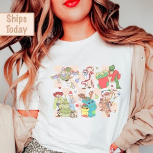 Puede incluir: Camiseta blanca con un estampado gráfico colorido de personajes de Toy Story vestidos con atuendos navideños. Los personajes incluyen a Woody, Buzz Lightyear, Rex y otros.