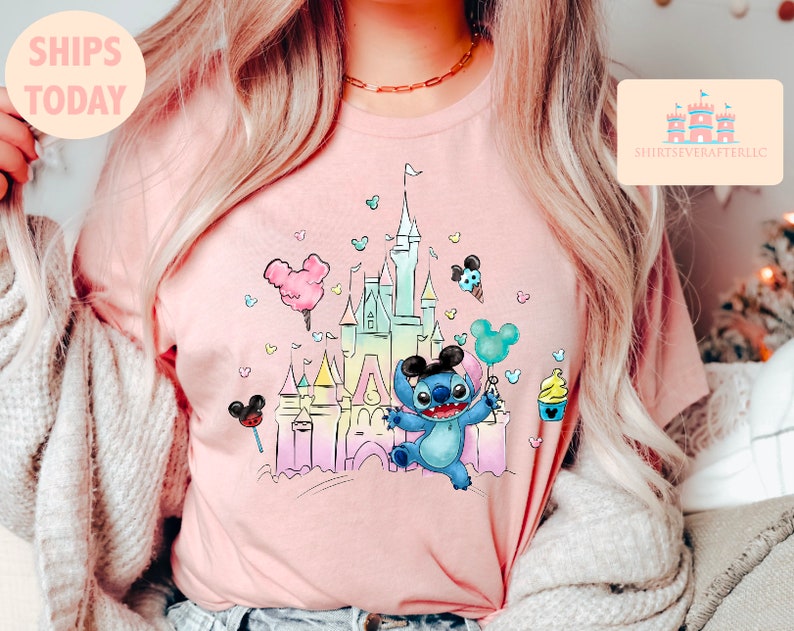 May include: A pink t-shirt with a colorful illustration of Stitch from Lilo & Stitch standing in front of a pastel-colored version of Cinderella's Castle. The illustration includes Mickey Mouse ears, a cotton candy balloon, and a Mickey Mouse ice cream cone.