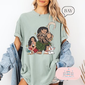 May include: A light green t-shirt with a graphic print of Maui, Moana, and Pua from the Disney movie Moana. The graphic is in a cartoon style and features Maui with his fish hook, Moana with her hair in a braid, and Pua the pig.