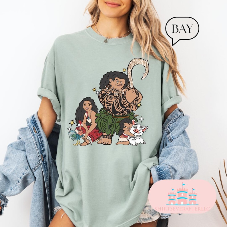May include: A light green t-shirt with a graphic print of Maui, Moana, and Pua from the Disney movie Moana. The graphic is in a cartoon style and features Maui with his fish hook, Moana with her hair in a braid, and Pua the pig.