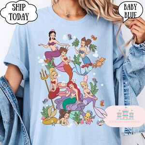 Op de afbeelding: Babyblauwe t-shirt met een kleurrijke illustratie van zeemeerminnen met verschillende haarkleuren en zeemeerminstaarten. Het shirt heeft de teksten "SHIP TODAY" en "BABY BLUE" in tekstballonnen. Het shirt heeft ook de tekst "SHIRTSEVERAFTERLLC".