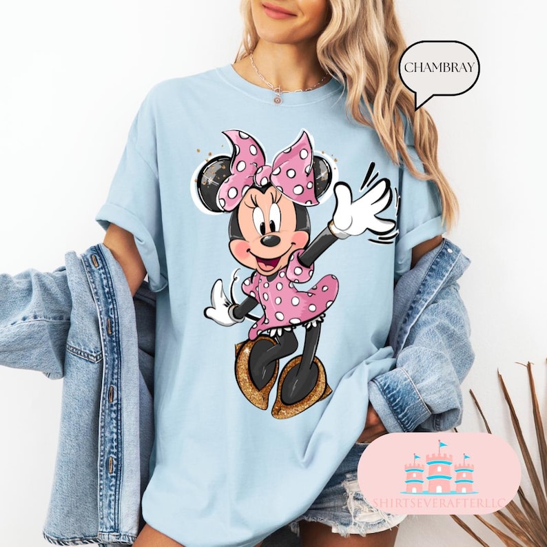Puede incluir: Una camiseta azul claro con un gr&aacute;fico de Minnie Mouse con un vestido de lunares rosas y un lazo rosa. El texto "CHAMBRAY" est&aacute; en un bocadillo de di&aacute;logo sobre Minnie Mouse.