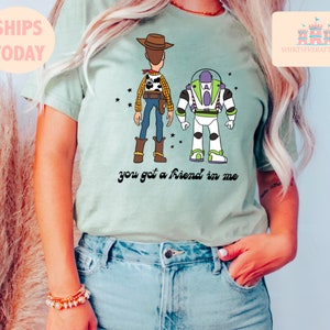 May include: A light green t-shirt featuring a cartoon image of Woody and Buzz Lightyear from Toy Story. The text "you got a friend in me" is printed below the characters.