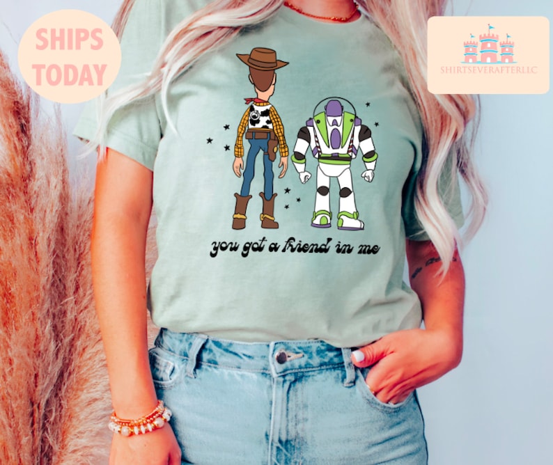 May include: A light green t-shirt featuring a cartoon image of Woody and Buzz Lightyear from Toy Story. The text "you got a friend in me" is printed below the characters.
