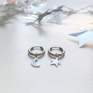 May include: A pair of silver hoop earrings. One earring features a crescent moon charm, and the other has a star charm. The earrings are displayed on a white surface with silver star decorations in the background.