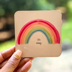 May include: A square, off-white coaster featuring a watercolor rainbow in shades of red, orange, yellow, green, and blue. The words "it's okay" are written below the rainbow in black script. The coaster is held in a hand.