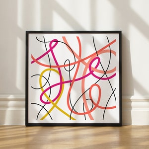 May include: Abstract art print in a black frame. The artwork features a white background with a colorful design of intertwined lines in shades of pink, coral, yellow, and black. The framed art is leaning against a wall.