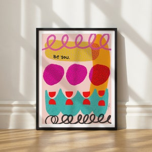 May include: A framed print with a pink background and colorful abstract shapes. The text "Be you." is in the center of the print.