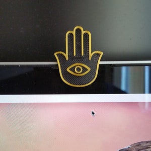 May include: A black hamsa hand with a gold outline and a gold eye in the center. The hamsa hand is a symbol of protection and good luck.