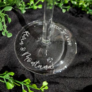 May include: Clear glass champagne flute with the names "Kelin & Montana" and the date "MAY 18, 2025" etched on the base. The glass is set against a black background with green foliage.