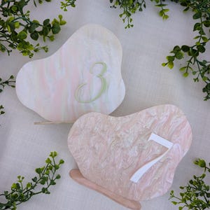 May include: Two heart-shaped table numbers with a marbled design. One is white and pink with a green number 3. The other is pink with a white number 7. Both have a light brown base and are surrounded by greenery.