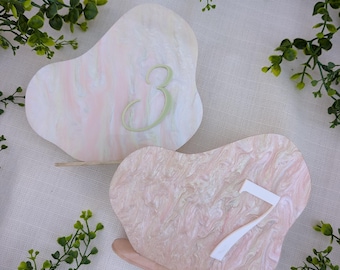 May include: Two irregular, pebble-shaped table number signs with a marbled finish in shades of pink, white, and cream. One sign features a light green number 3, while the other displays a white number 7, both supported by small stands.