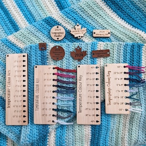 Peut inclure: Une couverture crochetée à rayures bleues, blanches et vertes, avec des clés de couleurs de température et des étiquettes en bois. Les étiquettes indiquent "2024 Temperature Blanket" et "Winnipeg, MB". Les clés de couleurs affichent les plages de température.