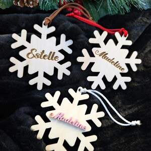 May include: Three wooden snowflake ornaments with the names Estelle, Madeline, and Madeline written on them. The ornaments are hanging from a brown, white, and red string.